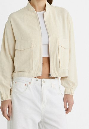 Beige cropped jakke med for-lynlås og klaplommer over hvid cropped top og højtaljede hvide bukser.