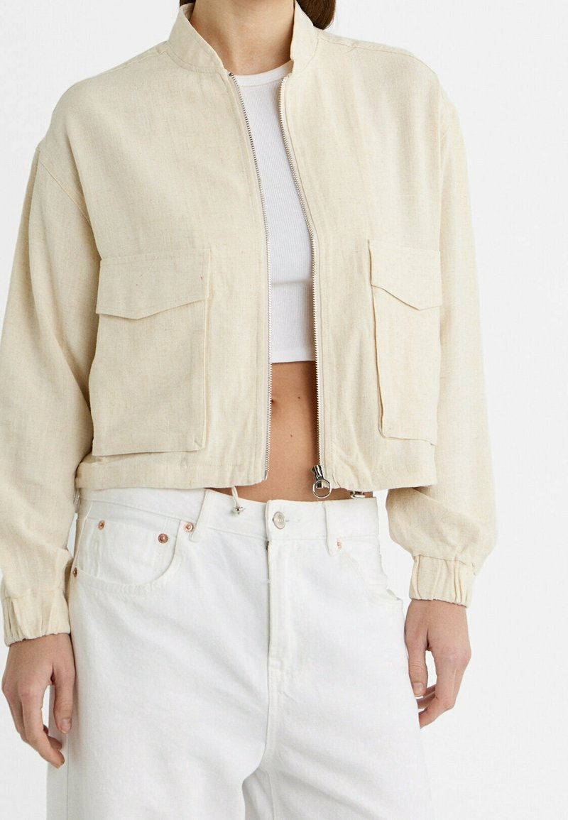 Veste courte beige avec fermeture éclair devant et poches à rabat portée sur un top court blanc et un pantalon blanc taille haute.