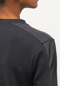 Sudadera negra con tejido texturizado en el hombro y detalles perforados, contrastando con un acabado liso en el cuerpo.