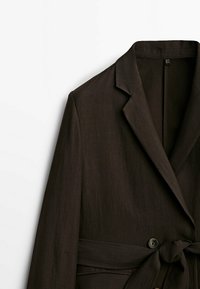 Blazer marron en tissu texturé, avec col à revers, ceinture nouée à la taille et fermeture par un bouton unique.