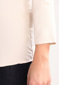 Blusa beige chiaro con una texture liscia, maniche 3/4 e cuciture laterali. Abbinamento suggerito con denim scuro per un contrasto.