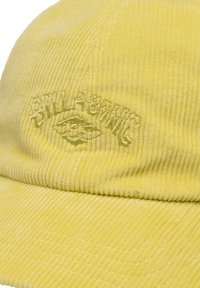 Gorra de pana amarilla con logo bordado. Presenta una textura acanalada y visera curva. Costuras de acento visibles a lo largo de los bordes.