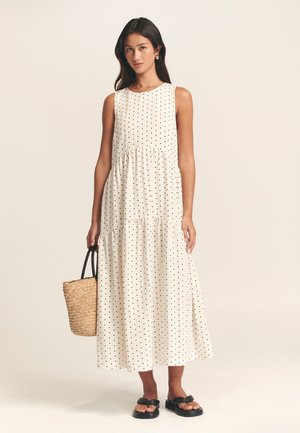 Krémové šaty bez rukávů s černými puntíky, vrstvený design a uvolněný střih. Doplněné tkanou slaměnou taškou a černými sandály.