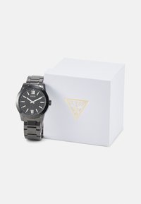 Schwarze Armbanduhr aus Edelstahl mit rundem Ziffernblatt, römischen Ziffern und glänzender Oberfläche. Begleitet von einer weißen, strukturierten Box mit goldenen Akzenten.