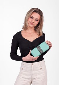 Young woman with shoulder-length blonde hair in black top and beige pants holding a turquoise wallet with a patterned strap against white background.