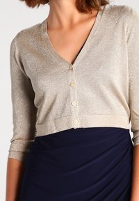 Short-sleeve, cropped cardigan in light gold, featuring a V-neck, five buttons, and a subtle shimmer, paired with a navy dress.
