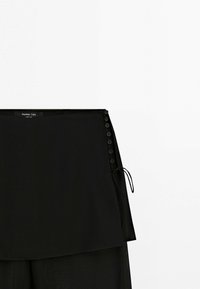 Jupe portefeuille noire avec fermeture à boutons sur le côté et lien réglable, étiquetée Massimo Dutti, présentée sur un fond blanc.