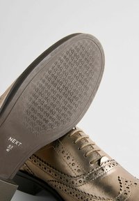 Chaussure habillée en cuir bronze avec des détails de brogue, texture lisse, bout rond et semelle en caoutchouc à motif. Informations de taille visibles sur la semelle.