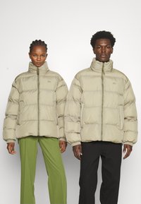Dos personas llevan chaquetas acolchadas de color beige claro con cuellos altos y cierres de cremallera. Las chaquetas cuentan con secciones acolchadas y bolsillos laterales.