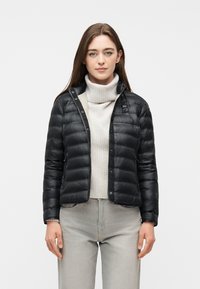 Femme portant une veste matelassée noire sur un pull à col roulé blanc et un jean gris clair, debout devant un fond uni.
