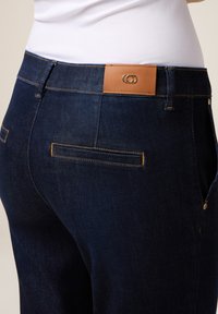 Jeans de denim de color azul oscuro, con un detalle en la cintura de cuero marrón claro y un corte ajustado. Incluye dos bolsillos traseros con acentos de costura.