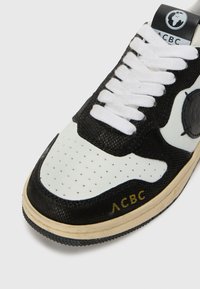 Čierno-biele tenisky s perforovanou špičkou, texturovanými materiálmi, bielymi šnúrkami, logom ACBC a žltým brandingom na boku.