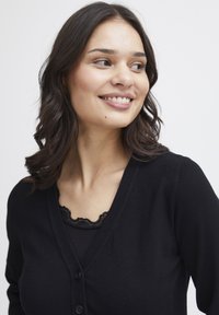 Svart stickad cardigan med djup V-ringning, som har ribbade ärmslut och knäppning med knappar. En tanktop med spetsdetaljer syns under.