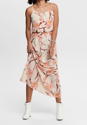 Kvinde iført en beige midi-kjole med pink blomsterprint, tynde stropper og hvide ankelstøvler, holder kjolen med den ene hånd.