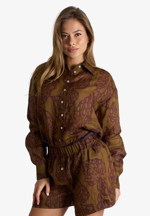 Donna che indossa una camicia marrone a maniche lunghe con bottoni e pantaloncini abbinati con grandi motivi floreali viola scuro, posando su sfondo bianco.