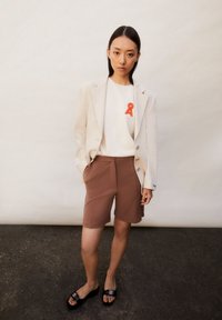 Lichtgrijze blazer over een wit T-shirt met een rode lintgrafiek, gecombineerd met bruine shorts. Zwarte sandalen met een dikke zool ronden de outfit af.