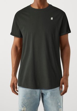 T-shirt nera di cotone con maniche corte arrotolate, colletto tondo e un piccolo logo sulla parte superiore sinistra. Indossato con jeans strappati di un blu chiaro.