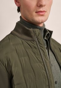 Green quilted jacket with a zip closure and ribbed collar, layered over a textured gray sweater. Smooth material with a subtle sheen.