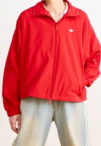 Czerwona kurtka na zamek wykonana z lekkiego materiału, z stojącym kołnierzem, elastycznymi mankietami oraz białym logo na lewej stronie klatki piersiowej. Casualowy design.