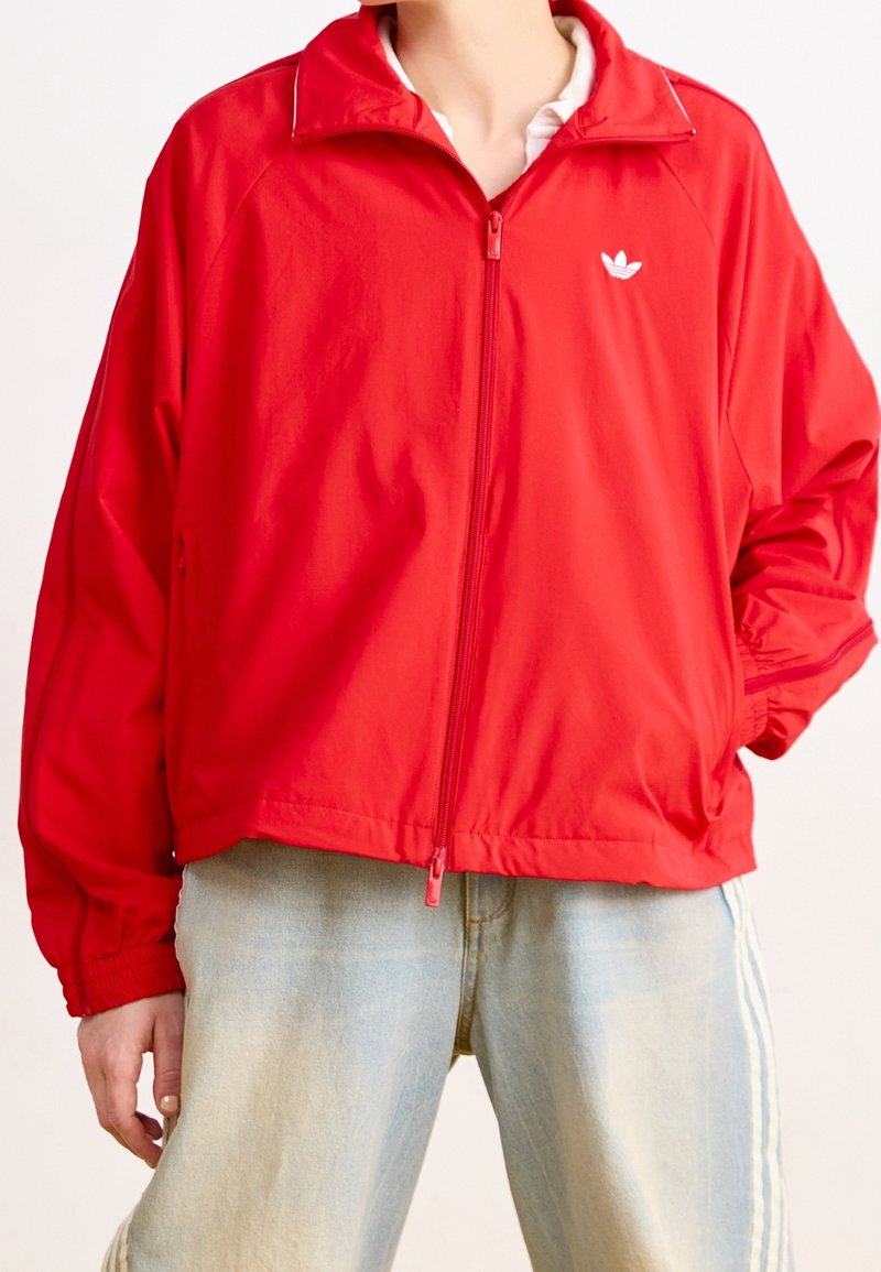 Czerwona kurtka na zamek wykonana z lekkiego materiału, z stojącym kołnierzem, elastycznymi mankietami oraz białym logo na lewej stronie klatki piersiowej. Casualowy design.