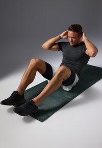 Schwarze Sport-Sneaker, graues enges Shirt, schwarze Shorts, führt einen Crunch auf einer grünen Trainingsmatte vor einem Farbverlaufshintergrund aus.