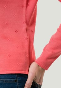 Suéter de punto coral con patrón floral texturizado, diseño ajustado y mangas largas. La tela parece suave, combinado con jeans de mezclilla oscuros.
