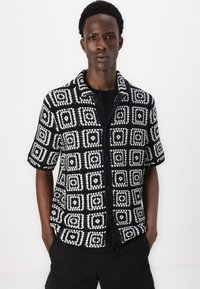 Chemise à manches courtes à boutons, en noir et blanc, avec des motifs carrés et des textures. Équipée d'un col et de boutons noirs. Tissu en mélange de coton.
