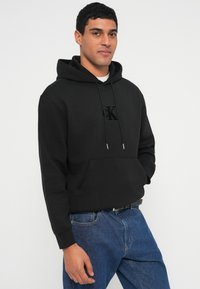 Camisola com capuz preta feita de material macio, apresenta um bolso frontal, capuz com cordão ajustável e um logotipo texturizado no peito. Usada com jeans azuis.