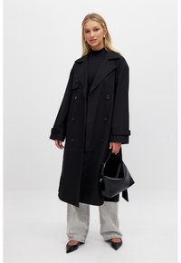 Cappotto lungo nero con design doppiopetto, ampi risvolti e polsini con cintura. Abbinato a jeans grigi e una borsa nera. Scarpe nere a punta.