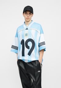 Ung mann som har på seg en svart caps, Argentina fotballdrakt med lyseblå og hvite striper med nummer 19, og svarte Adidas joggebukser.
