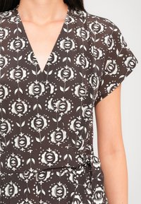 Femme portant une robe portefeuille marron à manches courtes avec un motif floral blanc nouée à la taille.