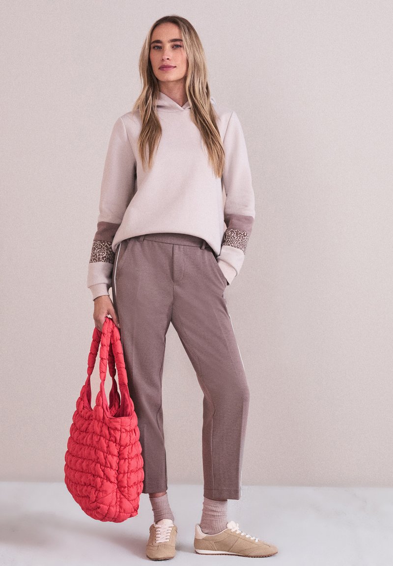 Femme portant un sweat à capuche beige et un pantalon mauve, tenant un grand sac fourre-tout matelassé rouge, debout devant un mur uni.