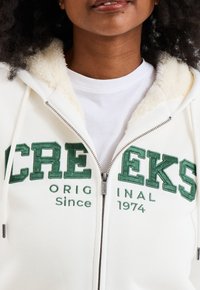 Witte rits-hoodie met een pluizige binnenkant, voorzien van groene geborduurde tekst: "CREEKS ORIGINAL Sinds 1974." Ritsdetail zichtbaar.