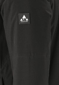 Schwarze wasserdichte Jacke mit glatter Textur, versehen mit einem rechteckigen weißen Logo-Patch mit der Aufschrift "WHISTLER" am Oberarm.