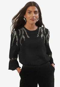 Blusa nera a maniche lunghe con intricati perline e un design floreale sulle spalle e volant a campana sui polsini. Texture liscia.