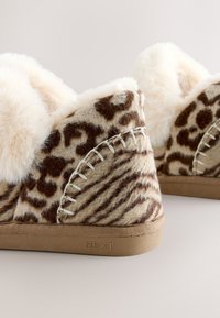 Chaussons à imprimé léopard avec doublure en fausse fourrure, tons bruns, texture douce, et détails de couture blanche sur une semelle en caoutchouc beige.
