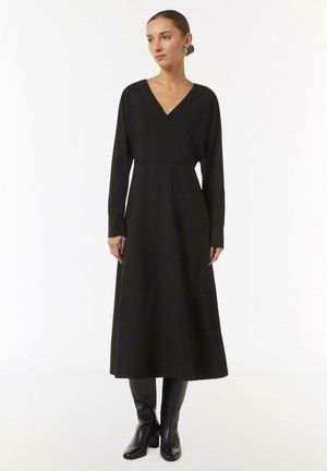 Schwarzes Langarmkleid mit V-Ausschnitt, tailliertem Oberteil, fließendem Rock und Säumen. Kombiniert mit knielangen schwarzen Lederstiefeln.