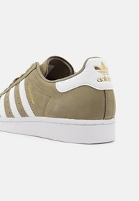 Beige Adidas Superstar-sneaker med vita ränder, vit hälflik, guld Adidas-logotyp och vit gummisula visad från en bakvinkel.