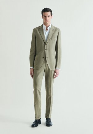 Hombre de pie, vestido con un traje verde oliva claro, camisa blanca y zapatos negros, frente a un fondo liso y claro.
