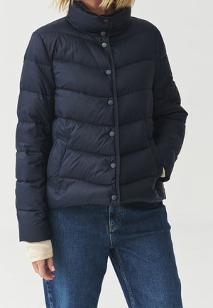 Vrouw draagt een marineblauwe gewatteerde donsjas met drukknopen en een spijkerbroek, met één hand in de jaszak en de andere zichtbaar.