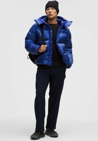 Sinine puhv jacket, millel on läikiv viimistlus, kapuuts ja tikitud muster, koos musta ülaosa ning tumesiniste kaubanduspüüksidega. Must beanie ja seljakott kuuluvad komplekti.