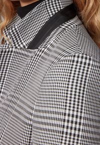 Tela de patrón de espiga en blanco y negro con un alto cuello y acentos en negro ribeteados contrastantes. Superficie texturizada, diseño angular.