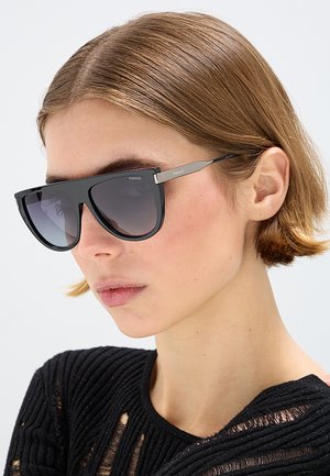 Giovane con capelli corti castano chiaro, occhiali da sole Polaroid neri e top nero strutturato su uno sfondo chiaro e uniforme.