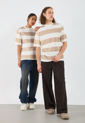 To modeller bærer matchende horisontale stribede t-shirts i beige og hvid, kombineret med løse jeans og afslappet fodtøj; baggrunden er helt hvid.