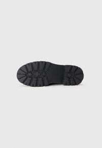 Semelle en caoutchouc noir avec des motifs de relief profonds, incluant des formes croisées et des rainures angulaires pour une adhérence et une durabilité optimales.