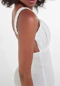 Bílé šaty se smocked texturou, širokými ramínky s nařasenými detaily a otevřeným zadním designem. Minimalistický styl s hladkým materiálem.