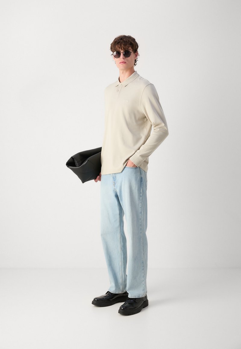 Polo shirt beige clair à manches longues, jean bleu détendu, chaussures noires et sac à main noir. Le mannequin porte des lunettes de soleil rondes.