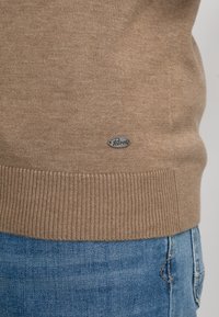 Ourlet de pull en maille marron avec petite étiquette ovale "Petrol" au-dessus de la ceinture d'un jean en denim bleu.