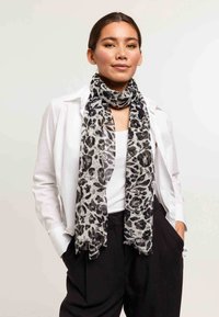Écharpe à motifs léopard en tissu doux et léger de couleur noire et grise, avec un bord effiloché, portée sur un haut blanc et un pantalon noir.