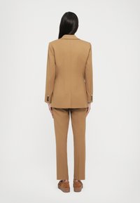 Brun skreddersydd dress med tilpasset blazer og rette bukser. Glatt tekstur, enkeltsidig design og knappdetaljer på ermene.